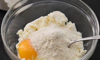 В миску выложите творог, если он зернистый, то разотрите при помощи вилки, а потом вбейте яйцо и всыпьте просеянную пшеничную муку
